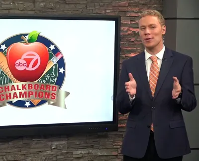 Male TV host standing beside screen displaying abc7 Chalkboard Champions logo with apple, ruler, and pencil