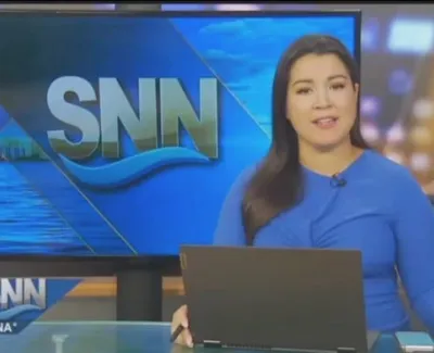 Female news anchor in blue dress presenting live on SNN news channel behind a laptop at 10:07 AM.