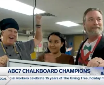 Three people celebrate ABC7 Chalkboard Champions in a classroom with a large check and smiles.