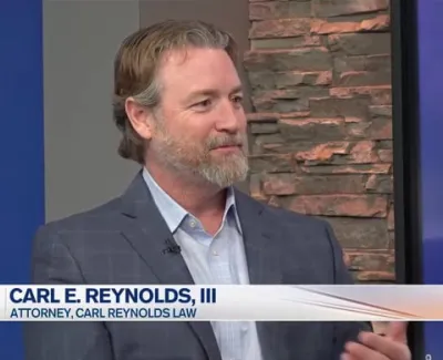 Attorney Carl E. Reynolds III speaking on TV news segment with stone wall background and blue studio decor