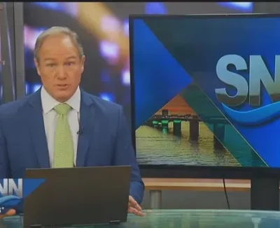 News anchor delivering evening edition on SNN with digital screen showing logo and bridge image.