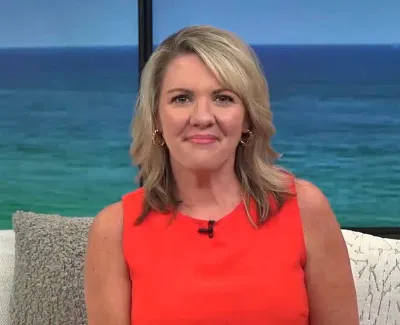 Smiling blonde woman in an orange sleeveless top sitting on a couch with ocean backdrop.