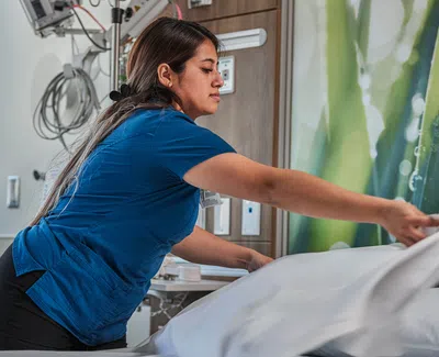 Healthcare worker in blue scrubs making a hospital bed with white linens in a patient room.