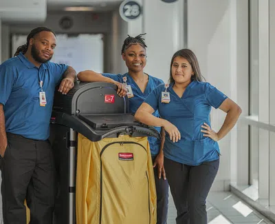 Three janitorial staff in blue uniforms pose with a yellow cleaning cart in a bright hallway.