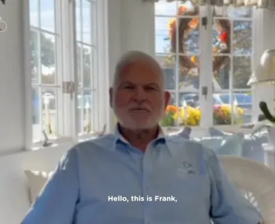 Older man named Frank sitting in a bright room with large windows and white furniture, wearing a light blue shirt.