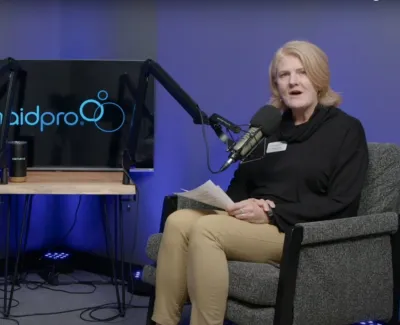 Two hosts sit in gray chairs with microphones recording a podcast in front of a MaidPro logo with purple blue lighting.