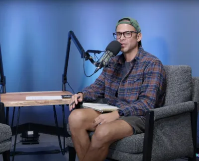 Two people recording a podcast in studio with microphones, seated on gray chairs against a blue backdrop.