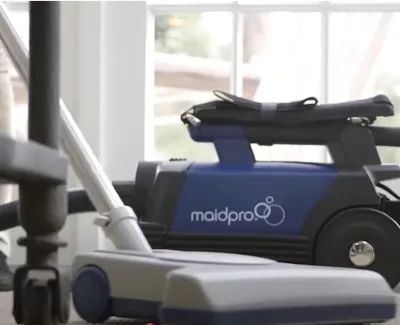 Maid Pro professional vacuum cleaner in use on carpet near office chair against glass door.