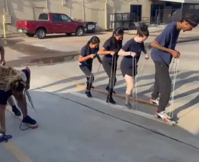 Group of young people outdoors practicing team-building activity using ropes on concrete ground under sunlight.