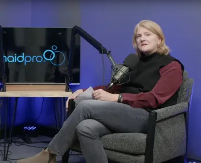 Two people seated in studio chairs recording a podcast with MaidPro logo displayed on a screen behind them.