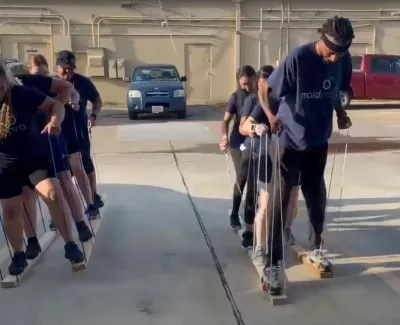 Teams participate in an outdoor team-building exercise using wooden stilts in a parking lot under sunlight.
