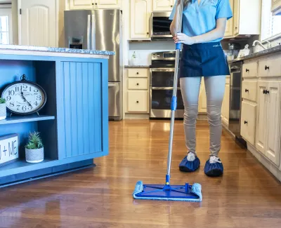 Person mopping hardwood kitchen floor wearing shoe covers with blue mop and modern appliances in background