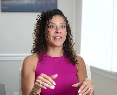 Woman with curly hair in a magenta sleeveless top speaking animatedly indoors by a window with white blinds.