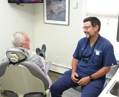 Dentist in navy scrubs smiling and talking with an older male patient seated in dental chair.