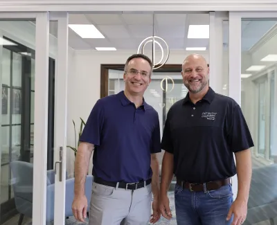 Two men smiling in casual polo shirts standing inside a modern office with glass doors and contemporary lighting.