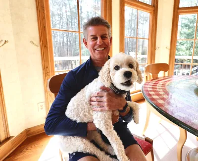 Man smiling and holding a white fluffy dog in a sunny dining room with large windows and a round table.