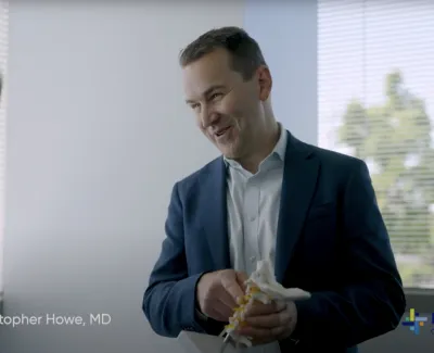 Doctor explaining spine model to female patient in a bright office with window blinds and greenery outside.