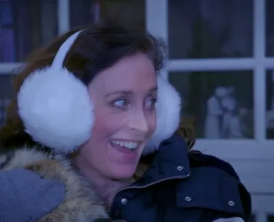 Woman wearing white fluffy earmuffs and winter coat smiling indoors by a window at night