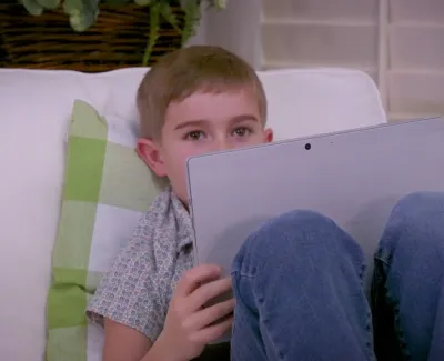 Young boy sitting on a couch holding a tablet with knees up near face in a cozy room.