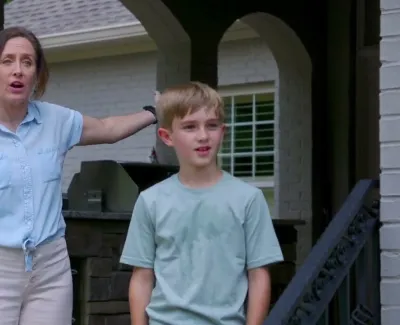 Woman talking with hand on hip beside young boy standing on porch with grill in background.