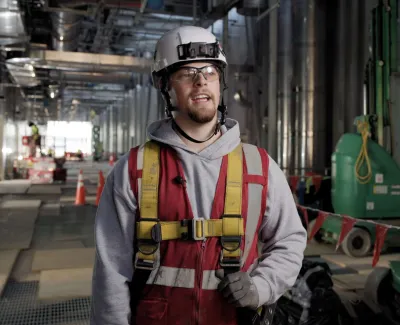 Construction worker wearing safety helmet, glasses, and harness stands inside an industrial building site.