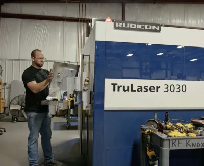 Man operating a large TruLaser 3030 CNC machine in an industrial workshop with tools nearby.