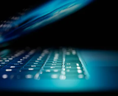 Close-up of a backlit laptop keyboard with glowing keys and partially open screen in low light.