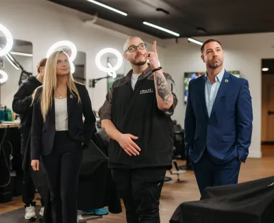 Three professionals stand inside a modern barbershop with ring lights and salon chairs visible in the background.