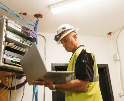 Technician in safety vest and helmet working on server rack using a laptop in a network equipment room.