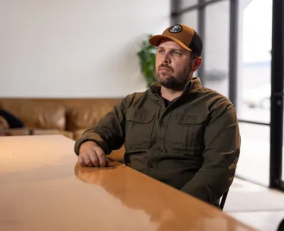 Man in a brown cap and green shirt sitting at a wooden table in a modern living room with a leather sofa.