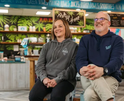 Two smiling adults in casual hoodies sitting inside a colorful cafe with tropical decor and menu boards.