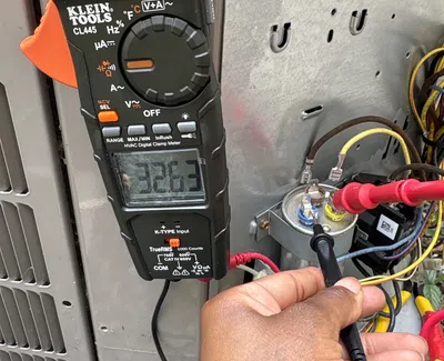 Hand holding black probe testing capacitor voltage with Klein Tools HVAC digital clamp meter showing 32.63 volts.