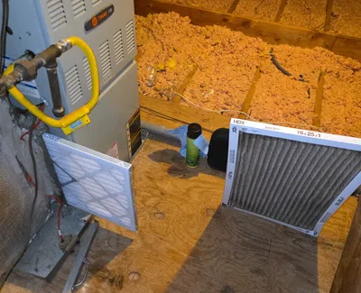Furnace system in an attic with new and dirty air filters on wooden floor near insulation material.