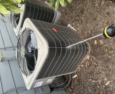Pressure washing an outdoor air conditioning unit with a hose on garden soil beside a house siding.