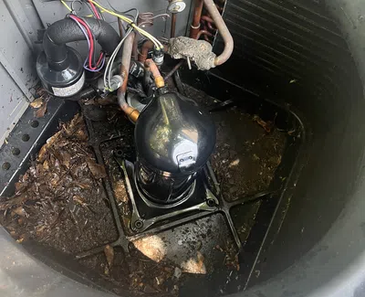 Interior view of a dirty HVAC unit showing compressor, copper pipes, wiring, and accumulated debris inside base.