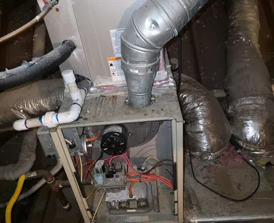 Open furnace unit in basement showing ductwork, pipes, wiring, and ventilation system components for heating.
