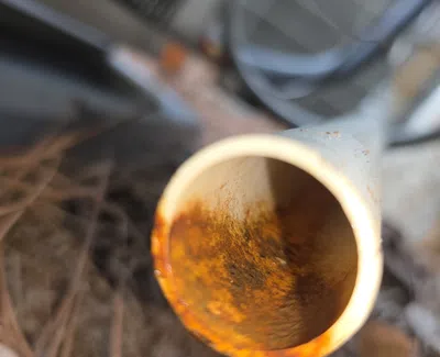 Close-up of a rusty metal pipe end with corrosion visible inside and pine needles on the ground.