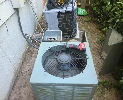 Two outdoor air conditioning units with repair tools and equipment in a narrow backyard area beside a house.