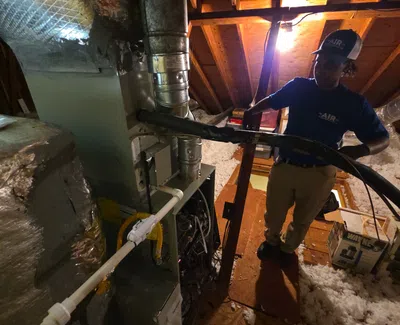 Technician inspecting and servicing HVAC system in attic with exposed wooden beams and insulation.