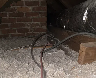Insulated attic space with ducts, wooden beams, brick wall, and electrical wiring over blown-in insulation.