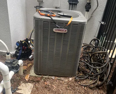 Outdoor Lennox air conditioning unit with tools, coiled hoses, and pipes against a textured white wall.