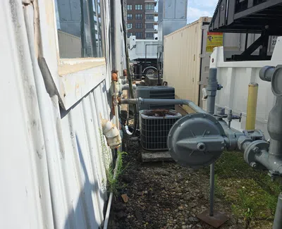 Outdoor industrial area with HVAC units, pipes, and gravel ground between buildings under a blue sky.