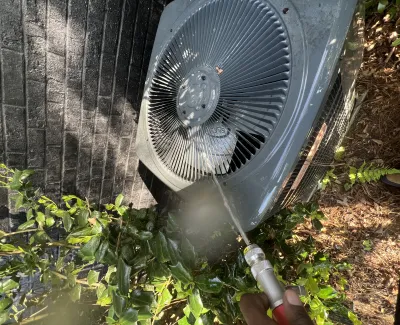 Person cleaning outdoor HVAC unit with a water hose surrounded by green plants and brick wall.