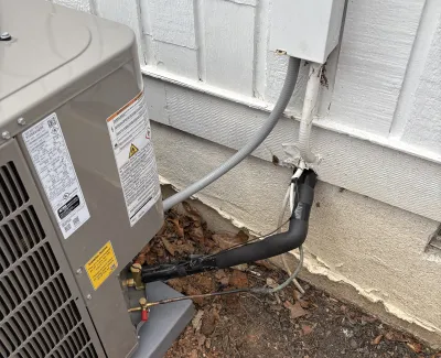 Outdoor air conditioning unit connected to a white wooden house with insulated pipes and electrical wiring.
