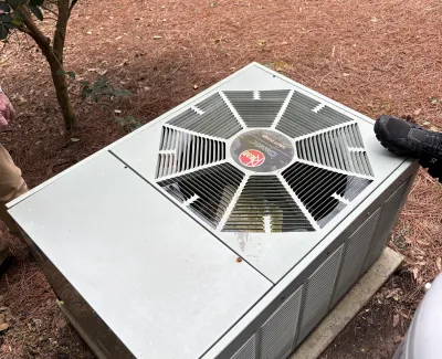 Outdoor rooftop air conditioning unit with fan grill and surrounding pine needles on ground in wooded area.