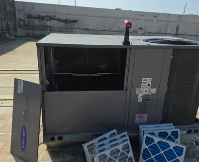 Open rooftop HVAC unit with removed air filters and a red cordless drill on top under clear sky.