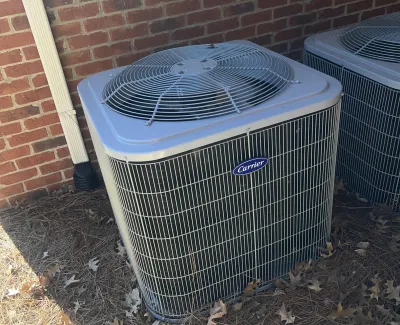 Outdoor Carrier brand air conditioning unit installed beside a brick house with dry leaves on the ground
