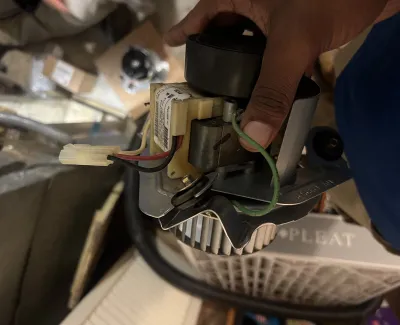 Hand holding electrical HVAC component with wires and metal parts in an indoor utility space.