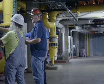 Two industrial workers wearing helmets operate machinery controls in a utility room with pipes and equipment.