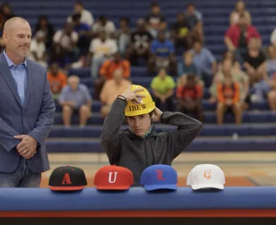 Young athlete wearing a yellow hard hat signing during a sports event with a man and colorful hats on the table